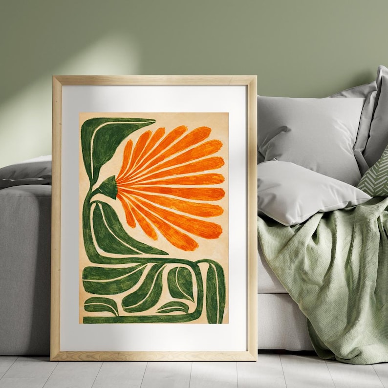 May include: A framed art print featuring a stylized orange flower with green leaves and stem. The artwork is set against a beige background and framed in a light wood frame. The floral design is a bold, modern interpretation.