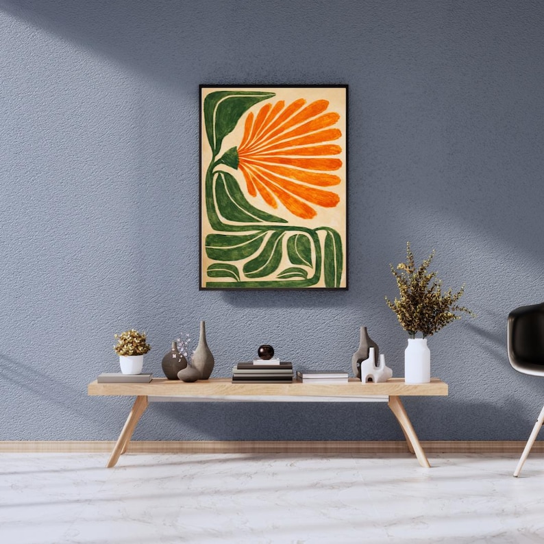 May include: Abstract floral art print in a black frame. The artwork features a large orange flower with green leaves and stems on a beige background. The print is displayed on a blue wall above a wooden table with decorative items.