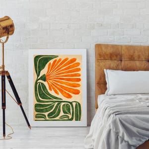 May include: An abstract art print featuring a stylized orange flower with green leaves and stems. The artwork is framed in a white border and leans against a white brick wall. The color palette includes orange, green, and cream.