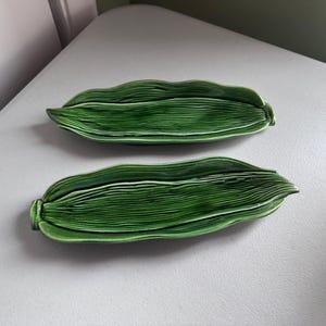 Puede incluir: Dos platos verdes de cerámica con forma de hoja. Los platos tienen un diseño texturizado y en capas, con un acabado brillante. Los platos están colocados sobre una superficie de color claro. El tema general es la vajilla decorativa.