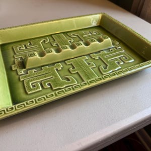 May include: Green ceramic ashtray with a rectangular shape and a raised, decorative design. The ashtray features a series of stylized, geometric patterns and grooves for resting cigarettes. The edges are adorned with a repeating scroll pattern.
