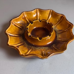 May include: A vintage ceramic ashtray with a unique, scalloped edge design. The ashtray features a central circular area for holding cigarettes, surrounded by a wide, petal-shaped rim. The color is a blend of brown and golden yellow.