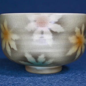 Puede incluir: Un cuenco de cerámica con un esmalte gris claro y un diseño floral. Las flores son blancas con centros amarillos y naranjas. El cuenco tiene forma redondeada y un pie pequeño.