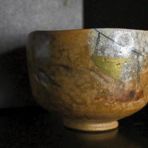 May include: A handcrafted ceramic tea bowl with a unique, textured surface. The bowl features a blend of brown, gold, and white hues, with gold leaf accents and black line details. The tea bowl is a traditional Japanese style.