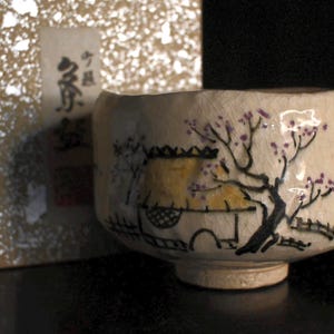Puede incluir: Un cuenco de té de cerámica color crema con una escena de paisaje pintada a mano que presenta una casa, un árbol y flores moradas. El cuenco está junto a una caja dorada texturizada con texto japonés.