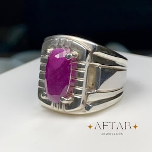 May include: A silver ring featuring a central oval-cut faceted deep pink ruby gemstone held by four prongs. The band has a wide, structured design with vertical grooved detailing around the stone. The text AFTAB JEWELLERS is visible.