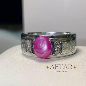 May include: A silver-toned ring featuring a central oval cabochon-cut pink gemstone, flanked by two small vertical rows of clear stones set in textured metal bands. The text AFTAB JEWELLERS is printed in the lower right corner of the image.