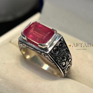 May include: A silver ring with a rectangular, red gemstone. The ring features intricate, dark floral engravings on the band and the text "AFTAB JEWELLERS" in gold. The gemstone is set in a silver bezel.