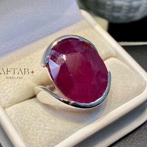May include: A silver ring featuring a large, oval-shaped, faceted deep red ruby gemstone set in a smooth metal band. The text AFTAB JEWELLERS is printed in gold on the left side of the white ring box interior, accompanied by two small star icons.