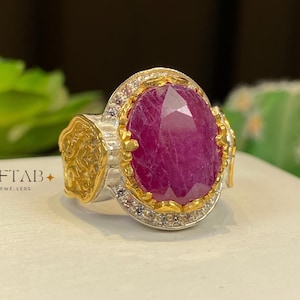 May include: A ring featuring a large, oval-cut, deep pinkish-red gemstone set in a gold-toned bezel. The band is silver-toned with gold-toned side accents and a halo of small clear stones. The text AFTAB JEWELLERS is printed on the left side.