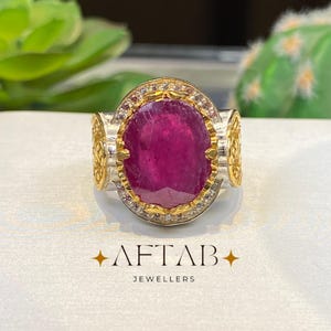 May include: Aftab Jewellers ring featuring a large, oval-cut ruby gemstone. The ruby is set in a gold and silver band with intricate detailing and small accent stones. The ring is displayed on a white surface.