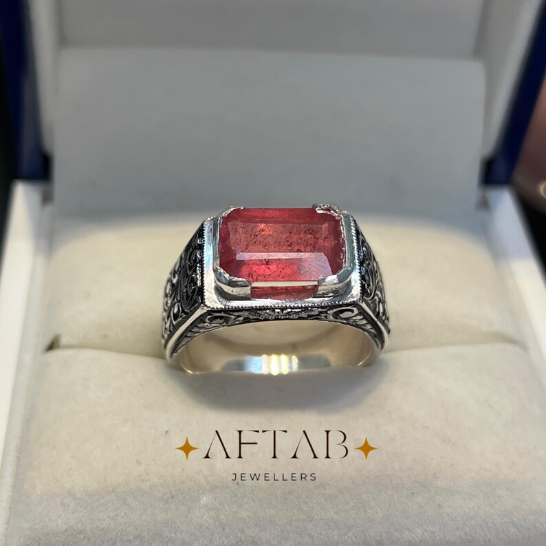 May include: A silver ring with a large, rectangular, red gemstone. The ring has an ornate, engraved design around the band. The text "AFTAB JEWELLERS" is at the bottom of the image.