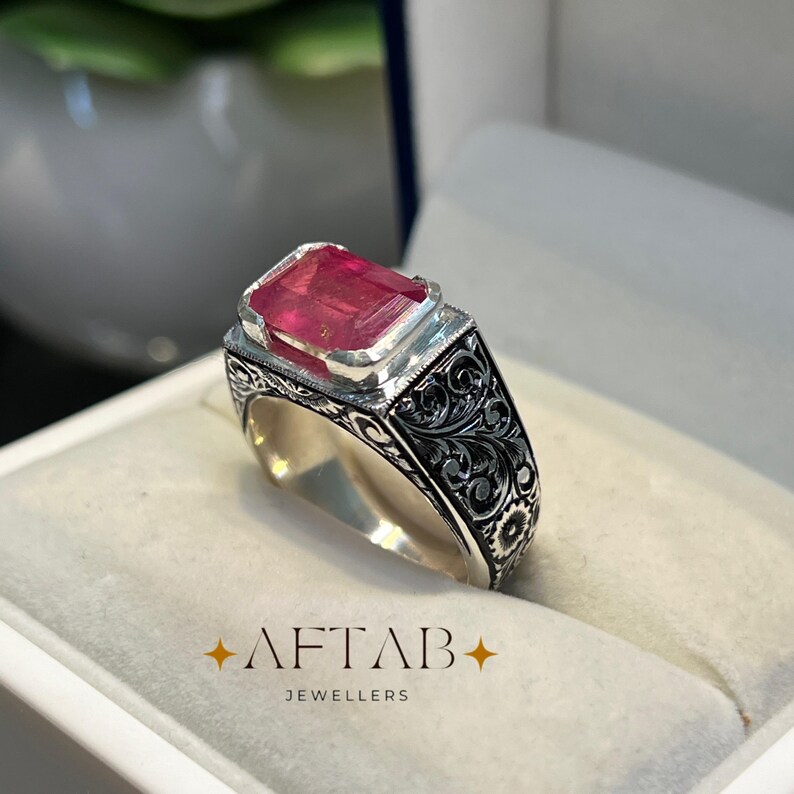 May include: A silver ring with a large, rectangular, red gemstone. The ring has intricate black floral engravings on the band and a square setting for the gemstone. The text "AFTAB JEWELLERS" is at the bottom.