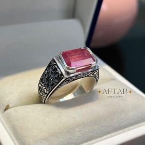 May include: A silver ring with an ornate, engraved band and a large, rectangular, pink gemstone. The ring is displayed in a jewelry box. The text "AFTAB JEWELLERS" is visible.