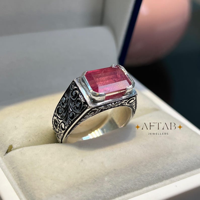 May include: A silver ring with an ornate, engraved band and a large, rectangular, pink gemstone. The ring is displayed in a jewelry box. The text "AFTAB JEWELLERS" is visible.