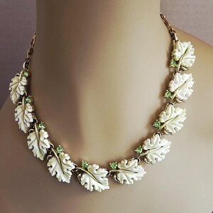 May include: A vintage necklace featuring cream-colored leaf-shaped pendants with gold-tone accents and small green gemstone embellishments. The necklace is displayed on a mannequin, showcasing its design and style.