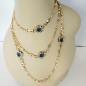 May include: Three-strand gold-tone necklace with blue circular accents. The necklace features a delicate chain design with decorative gold-tone elements between the blue circular stones. The necklace is displayed on a mannequin.