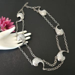 May include: A silver-toned necklace featuring a series of textured, curved pendant charms. The necklace has a double-strand chain design. The charms have a scale-like texture. The necklace is displayed on a white hand against a black background.
