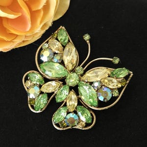 May include: A vintage butterfly brooch with a gold-tone metal frame. The brooch is adorned with various shades of green and yellow rhinestones, including oval, round, and marquise shapes. The wings and body are detailed with the sparkling stones.