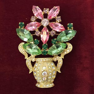May include: A gold-toned brooch in the shape of a vase overflowing with colorful gemstones. The brooch features pink and green crystal flower and leaf designs, with smaller green and purple accents. The vase is encrusted with clear crystals.