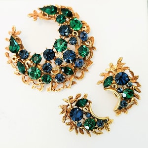 May include: A vintage jewelry set featuring a crescent-shaped brooch and matching earrings. The set is gold-toned with emerald green and sapphire blue rhinestones. The brooch measures approximately 3 inches in diameter.