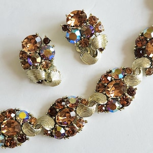 May include: A jewelry set consisting of a bracelet and a pair of clip-on earrings. Each piece features gold-tone metal with a brushed texture, accented by clusters of round and oval faceted brown crystals and iridescent blue-tinted stones.