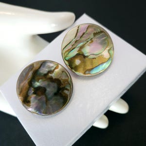 May include: A pair of round iridescent earrings made from abalone shell. The earrings have a brown, green, and purple color scheme.