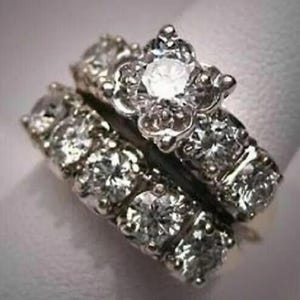 Puede incluir: Dos anillos de plata con piedras preciosas claras. Uno presenta un diseño floral con una piedra central grande. El otro anillo tiene una fila de piedras más pequeñas. Los anillos están sobre un fondo blanco suave.