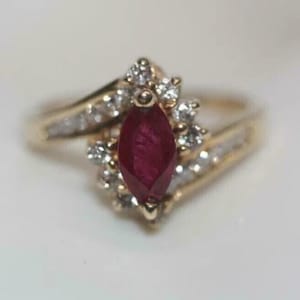 May include: A gold ring featuring a central marquise-cut deep red ruby gemstone. The ruby is surrounded by a cluster of small, clear, round-cut white diamonds set in gold. The band is also gold and includes additional small diamonds along the sides.