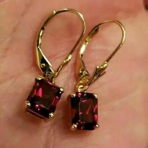 May include: A pair of gold-colored leverback earrings, each featuring a rectangular, deep red gemstone. The earrings are displayed against a soft, warm-toned background, highlighting the contrast between the metal and the stones.