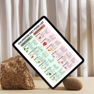 May include: A digital tablet displays a "Galveston Diet Food List" with foods to enjoy and avoid. The tablet is propped up by a large, textured rock and a small, smooth stone. The background features a white curtain.