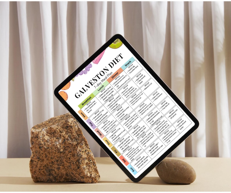 以下が含まれることがあります： 「GALVESTON DIET」と題された7日間の食事プランが表示されたデジタルタブレット。プランは日と食事ごとに整理され、朝食、昼食、夕食、軽食の列があります。タブレットは岩と小さな石で支えられています。