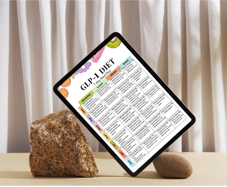 May include: A digital tablet displays a colorful 7-day meal plan labeled "GLP-1 DIET." The plan includes sections for breakfast, lunch, dinner, and snacks, with calorie and nutritional information. The tablet is propped up by a rock, with a neutral background.