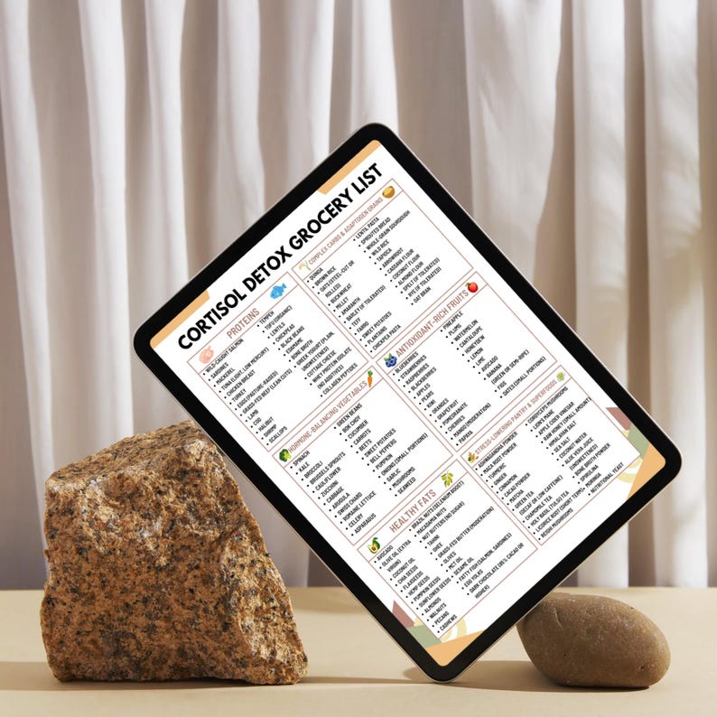 Puede incluir: Una tableta digital que muestra una "Lista de compras de desintoxicaci&oacute;n de cortisol" con categor&iacute;as como Prote&iacute;nas, Verduras que equilibran las hormonas y Grasas saludables. La tableta est&aacute; apoyada por una roca y una piedra peque&ntilde;a.