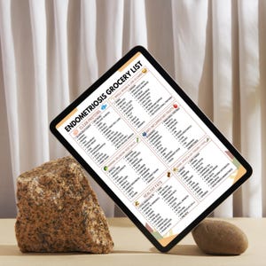 May include: A digital tablet displaying an "Endometriosis Grocery List" with categories like Clean Proteins, Anti-Inflammatory Fruits, and Healthy Fats. The tablet is angled, with a neutral background and two stones.