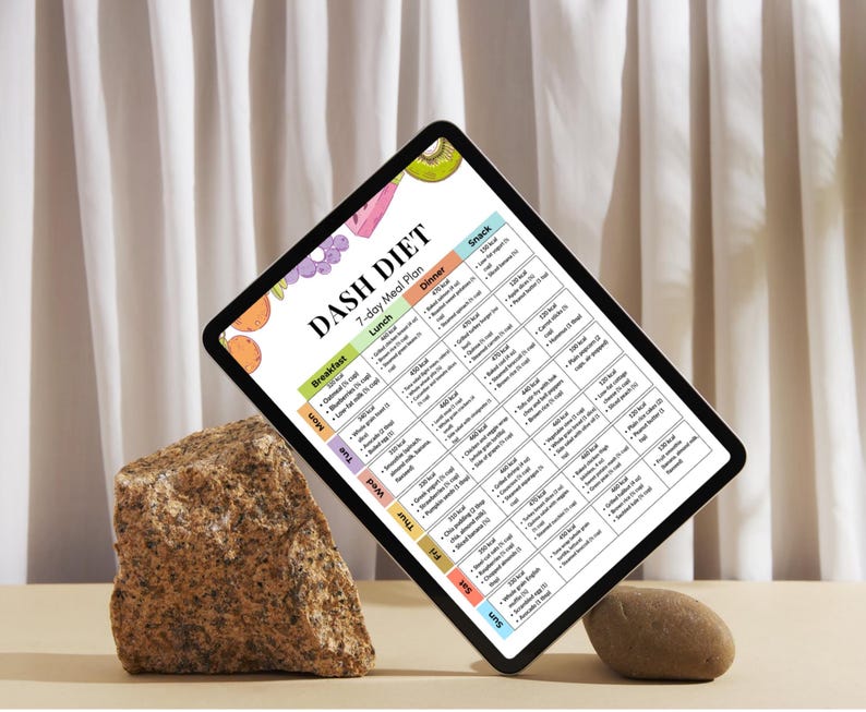 May include: A digital tablet showing a 7-day DASH diet meal plan. The tablet is supported by a rock and a small stone. The meal plan includes breakfast, lunch, dinner, and snack options with calorie counts. The tablet is angled, with a white curtain in the background.