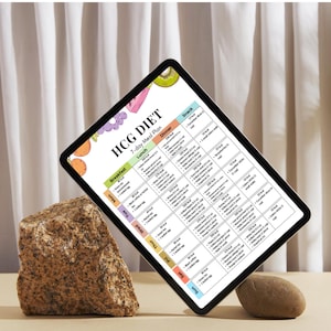 May include: A digital tablet displaying a 7-day meal plan labeled "HCG DIET." The plan includes breakfast, lunch, dinner, and snack options. The tablet is propped up by a rock and a small stone, set against a neutral backdrop.
