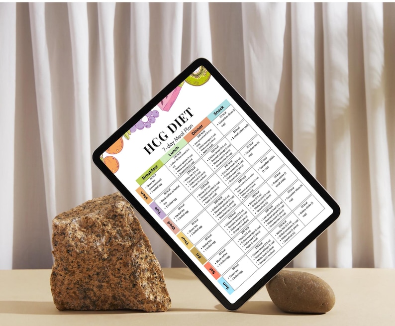 May include: A digital tablet displaying a 7-day meal plan labeled "HCG DIET." The plan includes breakfast, lunch, dinner, and snack options. The tablet is propped up by a rock and a small stone, set against a neutral backdrop.