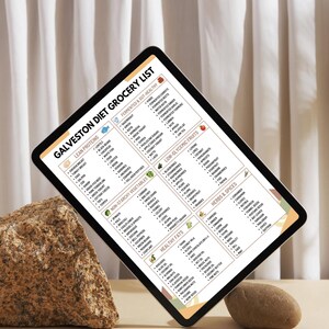 Może przedstawiać: Tablet cyfrowy wyświetlający "Galveston Diet Grocery List" z kategoriami takimi jak Chude białka, Warzywa nieskrobiowe i Zdrowe tłuszcze. Tablet jest podparty skałą i małym kamieniem na neutralnym tle.
