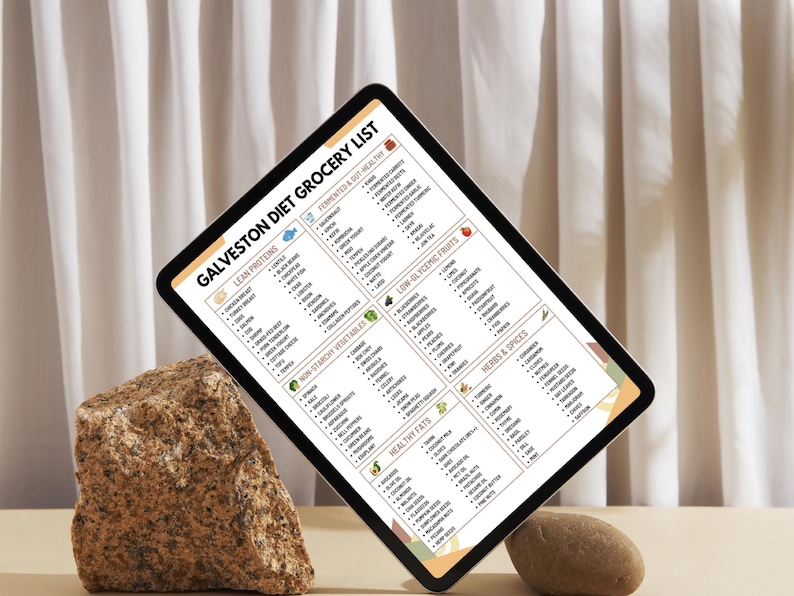 Może przedstawiać: Tablet cyfrowy wyświetlający "Galveston Diet Grocery List" z kategoriami takimi jak Chude białka, Warzywa nieskrobiowe i Zdrowe tłuszcze. Tablet jest podparty skałą i małym kamieniem na neutralnym tle.
