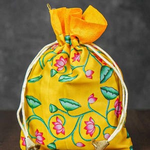 May include: A bright yellow drawstring pouch featuring a repeating pattern of pink lotus flowers and green lily pads on vines. The bag is cinched at the top with white cords that have metallic gold-toned bell-shaped ends at the bottom corners.