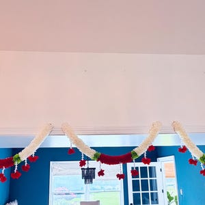May include: Three decorative garlands, each with a cream-colored, fluffy base and a vibrant red central band. Red floral accents and green leaves are interspersed along the garland, creating a festive and colorful display.