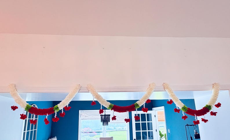 May include: Three decorative garlands, each with a cream-colored, fluffy base and a vibrant red central band. Red floral accents and green leaves are interspersed along the garland, creating a festive and colorful display.