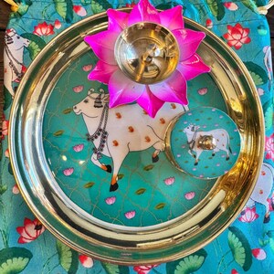 May include: A gold-toned metal plate featuring a central illustration of a white cow, accompanied by a matching small round container and a bright pink lotus-shaped metal oil lamp, all resting on a turquoise fabric pouch with a floral pattern.