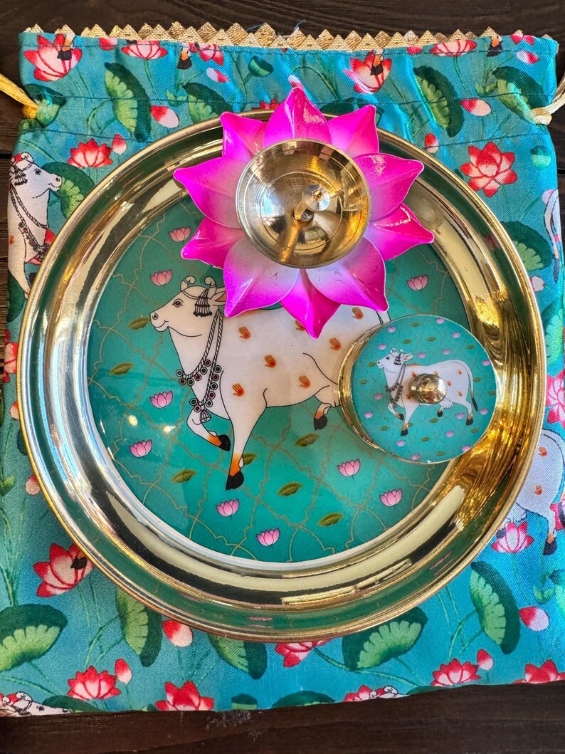 May include: A gold-toned metal plate featuring a central illustration of a white cow, accompanied by a matching small round container and a bright pink lotus-shaped metal oil lamp, all resting on a turquoise fabric pouch with a floral pattern.