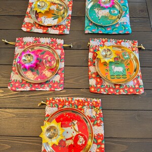 May include: Five sets of round gold-toned metal plates, each featuring a central illustration of a cow. Each plate includes a lotus-shaped metal diya lamp and a small round container, all resting on a drawstring fabric pouch with a cow print.