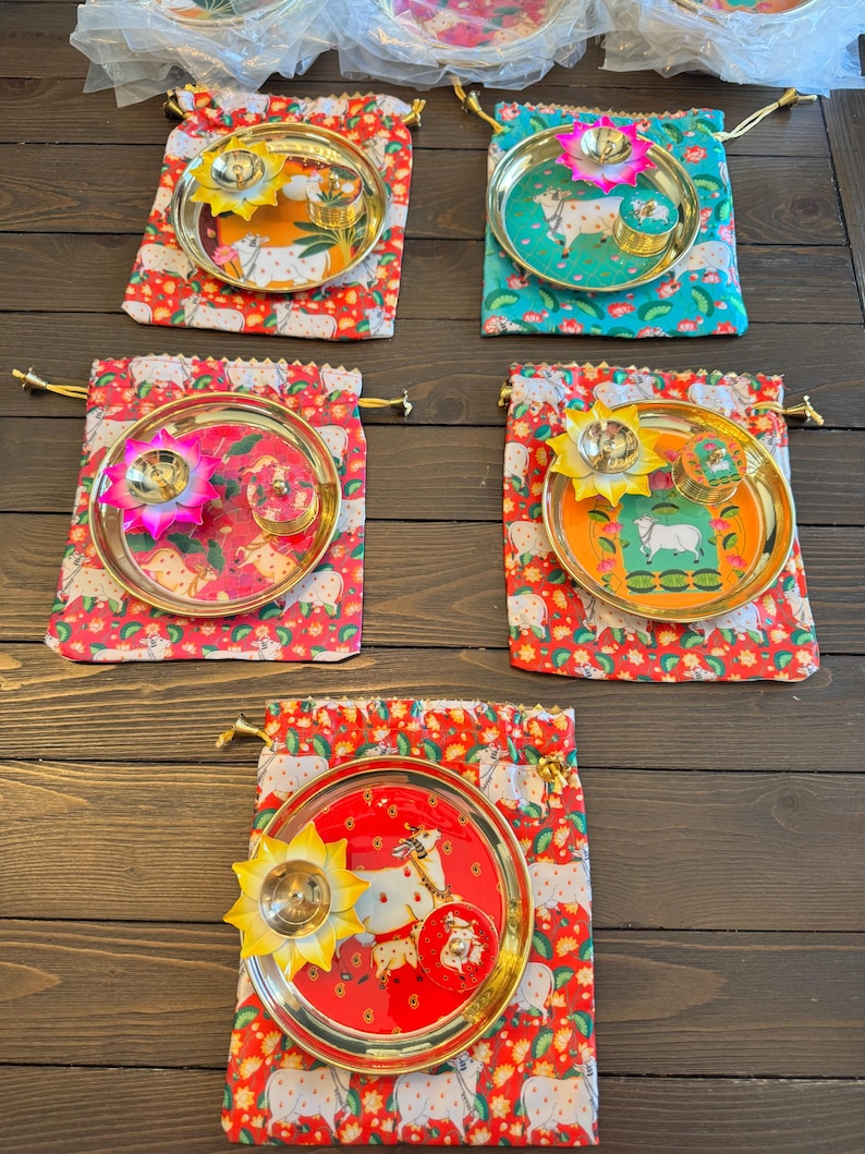 May include: Five sets of round gold-toned metal plates, each featuring a central illustration of a cow. Each plate includes a lotus-shaped metal diya lamp and a small round container, all resting on a drawstring fabric pouch with a cow print.
