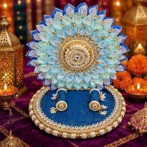Puede incluir: Un soporte de exhibici&oacute;n circular azul con una plataforma central elevada y un respaldo decorativo de tela azul y azul claro en forma de loto. La base est&aacute; adornada con bordes dorados y cuentas blancas, e incluye dos peque&ntilde;os soportes cil&iacute;ndricos.