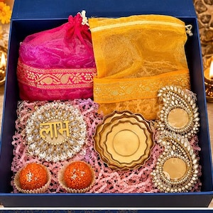 May include: A blue gift box containing a pink drawstring pouch, a yellow drawstring pouch, a circular decorative piece with the Hindi word Labh, two gold-toned metal lotus-shaped candle holders, and two orange round sweets in paper liners.