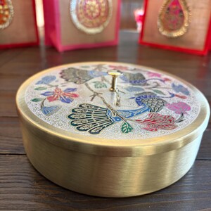 May include: A round, brass-colored container with a decorative lid featuring a peacock and floral design in blue, red, and green. The container sits on a dark wooden surface. In the background are pink and tan gift bags.